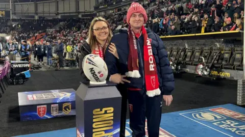 A man standing with the aid of his wife next to a plinth that has a match ball placed on top. Both have a red scarf around their necks and the man is wearing a woolly red hat. They are in a stadium with supporters in the background.