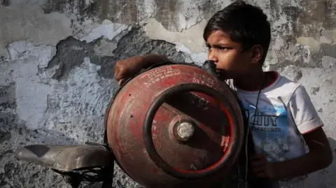 A boy stands next to an empty LPG cylinder tied to a bicycle as he waits outside a gas agency in India
