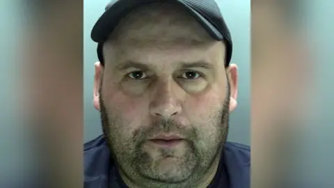 Merseyside Police Police custody image of Steven Naylor with dark brown stubble and brown eyes wearing a navy t-shirt and black baseball cap.