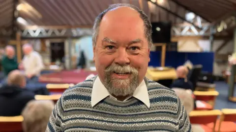 A man with grey hair and a goatee. he is looking directly at the camera whilst smiling. he has a grey and blue patterned jumper on with a white collar poking out.