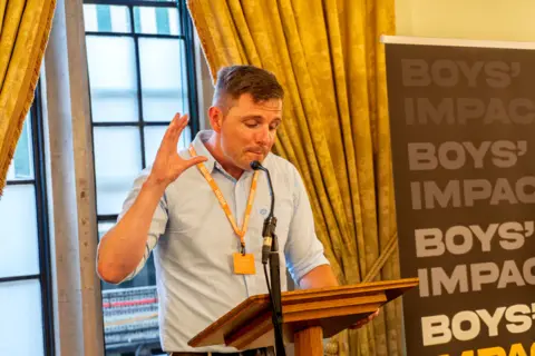 Supplied Dr Alex Blower is standing at a wooden lectern speaking into a microphone. He is wearing a blue checked shirt with his sleeves rolled up and is looking down at notes while he talks into the microphone. Behind him is a impact with the Boys' Impact logo. 