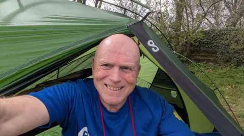 Alan Chape is smiling and sat inside the opening of his green tent. Woodland can be seen behind him. He is wearing a blue t-shirt.