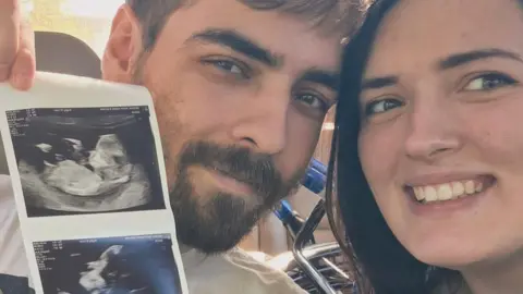 A man with short dark brown hair and a beard sits next to a woman with shoulder length brown hair on a hospital bed. He is holding up baby scan images to show the camera.