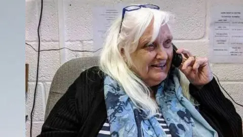Shaftesbury & District Carers Association A woman with white hair and blue glasses on her head. She is wearing a blue scarf and a black cardigan - she is using a black phone
