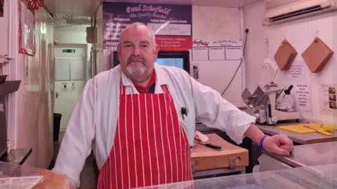 Butcher Paul Schofield stands behind his counter in The Quality Butcher on Market Street in Penistone. He is wearing a red and white striped butcher's apron. A yellow chopping board and knife are to the right of his hand, which is resting on the glass counter.