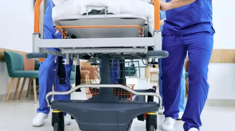 Hospital staff with royal blue scrubs and white shoes pushing a hospital bed which is metal with orange handles.