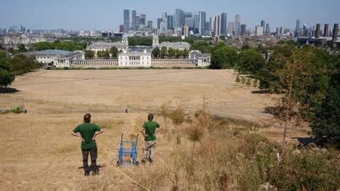 Getty Images Two council workers look out over Greenwich Park where the grass has turned brown. The historic buildings in Greenwich and the skyline of the City are in the background of the picture. 