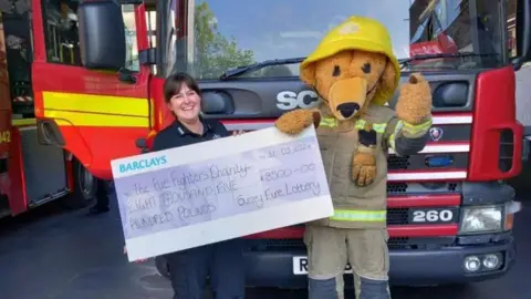 A member of the fire brigade and their mascot stands in front of a fire engine