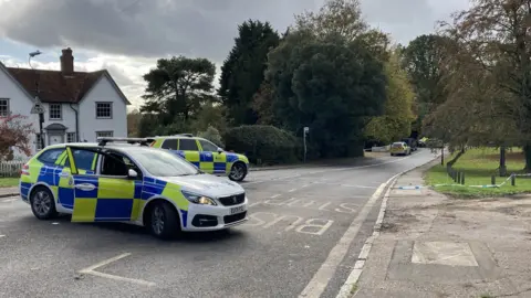 Stuart Woodward/BBC Two police cars blocking a road which is flanked by trees and a white house. Blue and white police tape is draped across the road too and in the background officers are investigating the crash scene.