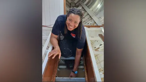 Contributed A woman works inside a boat hull during its build. She is crouched on her knees and leaning over on her hands. She has looked up to smile at the camera. She has short dark braids and is wearing a blue T-shirt and dark trousers that are covered in splats of white paint.