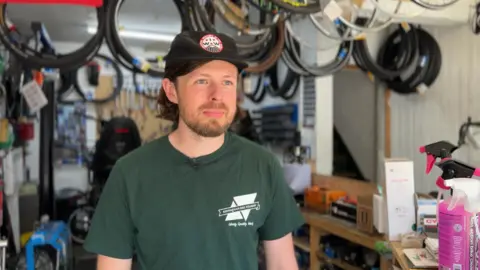 A man wearing a green t-shirt and a cap is standing in a room, with various bike tools and equipment behind him.
