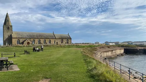 A church in the distance with an expanse of grass in the foreground and a sea wall to the right.