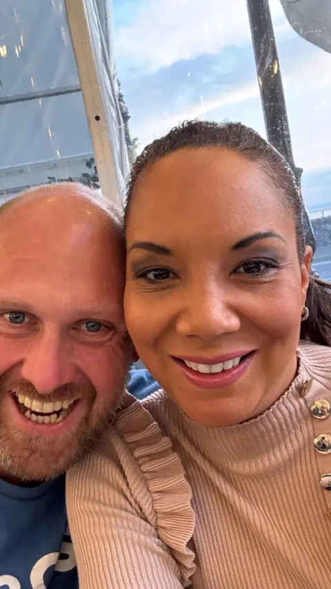 A man and women smiling at the camera. She has dark brown hair and he is bald. 