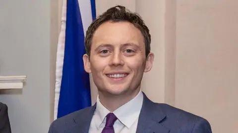 Mr Patrick has short brown hair and is smiling in the picture as he wears a grey blazer, purple tie and white shirt.