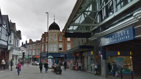 A general view of Wellingborough town centre, showing the entrance to the Swansgate Shopping Centre on the right and other stores on the pedestrianised street. There are shoppers walking about and people sat outside a coffee shop.