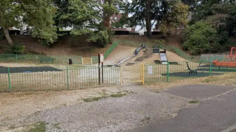 The image shows a playground that contains a slide, a seesaw and some swings. Behind the park are trees and some houses that can be seen behind them.