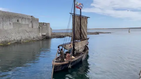 A traditional wood and leather sail boat with sails on a calm sea with a stone wall to the left as it sails towards the horizon.