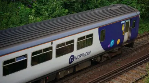 A Northern Trains carriage with its white and blue colours on a railway track with thick trees behind.