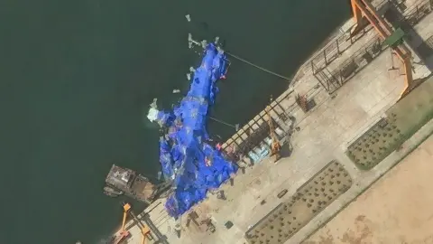 A satellite image shows the warship lying on its side, covered by large blue tarpaulins. A portion of the vessel appears to be on land.