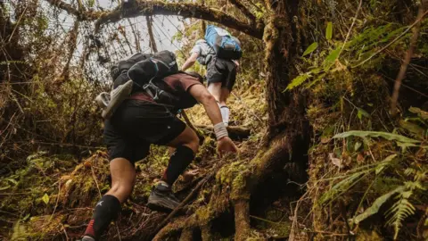 Jenny Scott Communications Two men are climbing in a steep forest area. Both are wearing black shorts and carrying bags on their backs.