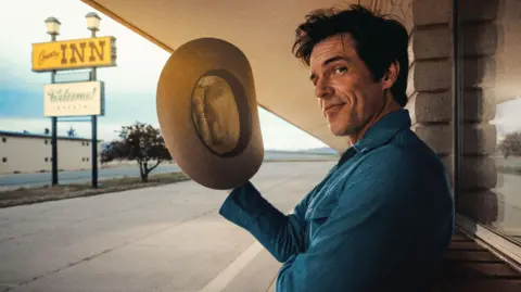Brandon Flowers holds a stetson in his right hand and gives a wry look to the camera as he poses outside a stereotypical American motel