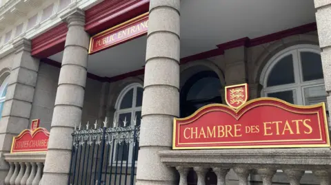 BBC The front and public entrance of Jersey's States Assembly. The stone-fronted building has an outdoor foyer before its front doors, with two pillars and a metal gates. It also has red signs with gold lettering saying "States Chamber" and "Chambre Des Etats".