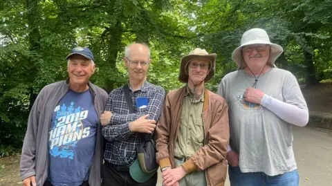 BBC/Sofía Luis-Hobbs Four men stand beside each other for a group photo on a woodland path. The man on the left wears a Leeds Rhinos navy cap and a navy tshirt under a grey jacket. Beside him a man with glasses wears a navy chequered shirt and has his arms folded across his chest and wears a cross body bag across him. To his right, a man wearing a khaki shirt tucked into his beige trousers. He also wears a brown bomber style jacket and a beige fishermen style hat. To his right, a man wearing a white bucket hat wears a grey tshirt over a long sleeved white top and has one of his arms resting on his chest.