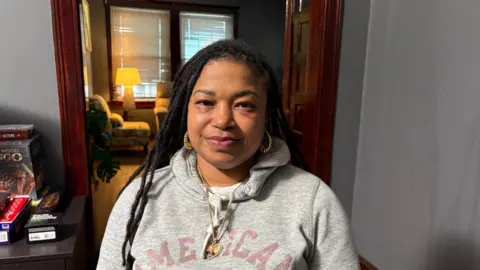 A woman with dark hair is standing in a doorway looking directly at the camera. She is wearing a grey sweatshirt which reads 'America' and is wearing large gold hoop earrings and a gold chain. She is standing beside a grey wall, with a brown wooden door and an armchair visible behind her. 