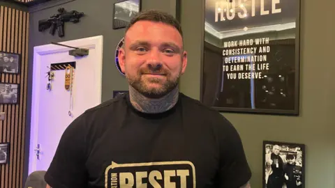 A man with short hair a beard and neck tattoo, wearing a black t-shirt. There are posters, pictures and a gun on the green wall behind him