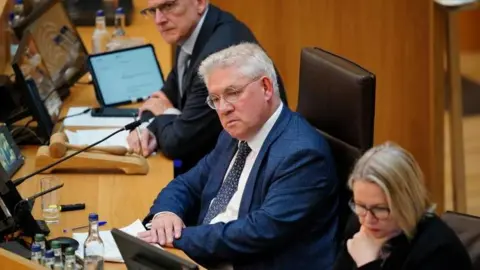 Kenneth Gibson, sitting at a desk with his hands sitting on top of each other. He is wearing a dark blue suit, and tie. There is a man and woman sitting at either side of him.