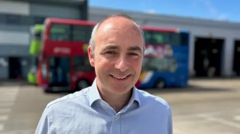 Ben is pictured at the Go North East bus depot in Gateshead, in front of two buses. He is wearing a blue shirt. He has short greying hair and black eyebrows.