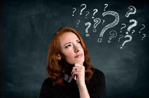 Picture of a woman with a quizzical expression standing in front of a blackboard with lots of question marks on it