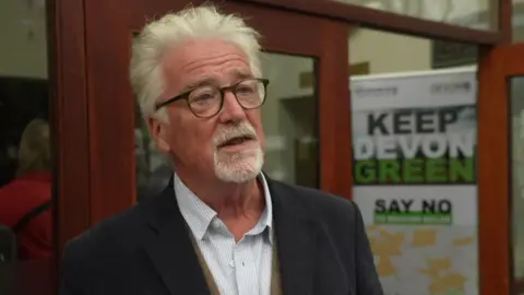 BBC A wide shot of Stephen Crowther. He has short grey hair, is wearing glasses, a white shirt, and a dark jacket.