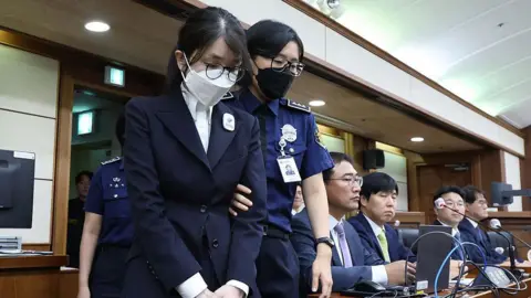 A woman in a black suit, face mask and glasses is escorted into a court by a police officer who is holding her by the arm. Four men in suits sit at a table behind them