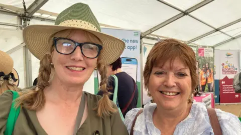 Vikki Irwin/BBC Two women stand together. The woman on the left has dark blonde hair that has been braided into two plaits on either side of her head. She wears a green and cream hat, black glasses and a green top. The other woman beside her is slightly shorter and has short red hair. She is wearing a blue and white blouse. 