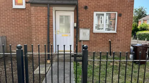 The front part of the house is on the Hartley road where the husband lived. A white front door and a black wall around the outside