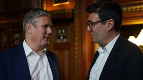 PA Media Andy Burnham and Keir Starmer in suits stand close together talking in an ornate, wood‑panelled room, with one holding a glass of water and warm lighting and framed decorations visible in the background.