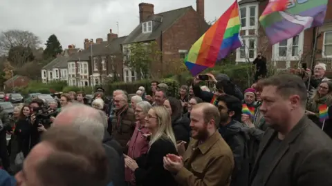 A large crowd of smiling men and women gather to watch the unveiling. They are standing in the road and there are houses visible behind them. Many are clapping, some are wearing colourful scarves and waving multi-coloured flags.