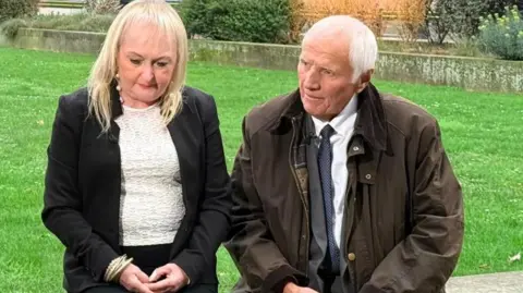 Tim Reid Media Jenni Hicks, who has blonde hair and is wearing a dark suit jacket over a white top, and Trevor Hicks, who has white hair and is wearing a grey suit and white shirt under a dark greenish brown overcoat, sit on a low wall in front of a patch of green grass and speak to a journalist who is behind the camera
