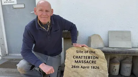 Stonemason Dave Greenhalgh, who is bald and wearing blue jeans and a blue sweatshirt, kneels beside a large stone that says "Site of the CHATTERTON MASSACRE 26th April 1826" in black text