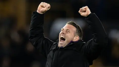 Rob Edwards raises his fists to the air and shouts in celebration, eyes closed