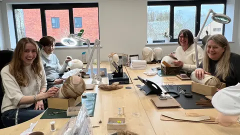 Four women sat around a wooden table hand-making wigs 