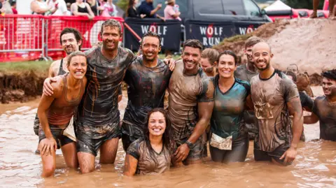 Nine people standing in a body of water that is brown and muddy. There are Three women and six men and have their arms around eachother. They are all covered head to toe in brown mud