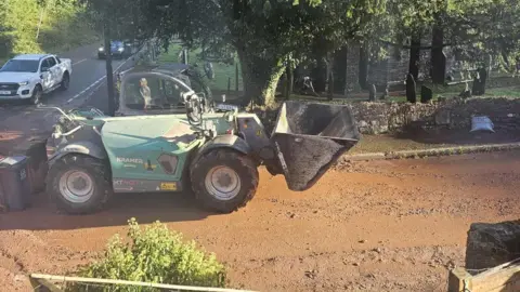 Rachel Jagger A large piece of machinery on a muddy road. It is green and there are cars travelling on the road behind. Behind the machinery are trees and a stone wall.