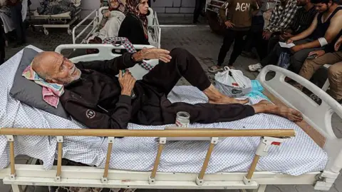 Getty Images An injured Palestinian man with a grey beard lies on a bed with wheels, with other injured people visible in the background