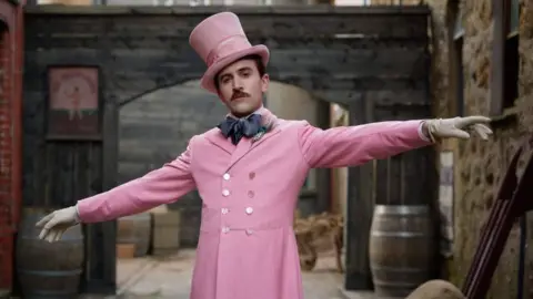 Callum Scott Howells dressed in one of Henry Paget's outfits. It is a pink buttoned up suit jacket paired with a large navy blue bow tie,. He is also wearing a pink top hat and has white gloves on his outstretched hands.