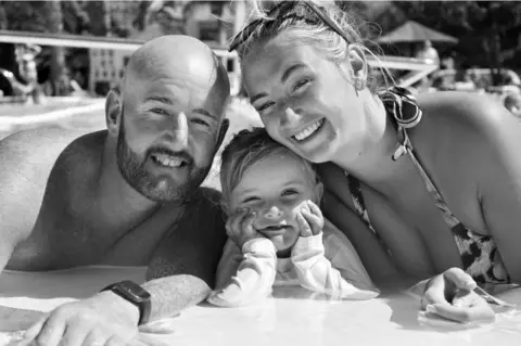 Warren Davies A black and white photo of a man and a woman with a youn girl between them. They are in a row, leaning up on the side of a swimming pool. The man is bald, with a beard, and is shirtless. The woman has ligth hair piled on her head with sunglasses perched on top. She is wearing a swimsuit. The little girls is resting her chin in her hands with her elbows propped up on the side. They are all smiling and looking at the camera.