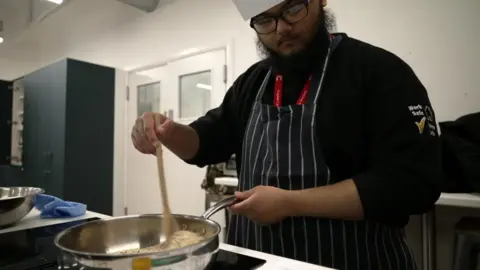 A young man in a chef's uniform stirs some ingredients in a saucean. He wears a white chef hat, a red lanyard, and a blue and white striped apron.
