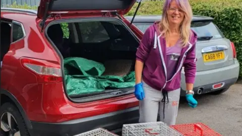 Anna Mason Anna Mason stood next to her car boot. She is wearing blue gloves and there are numerous cat cages in front of her.