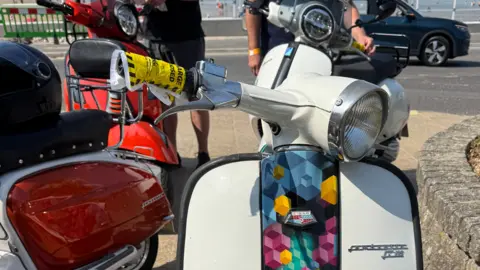 Close-up of a white scooter with a multicoloured decorative strip down the centre of the front faring and a yellow parking fine taped around one of the handlebars. Three other scooters are parked in the background.